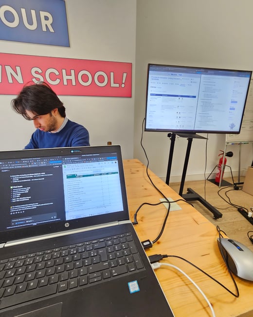 A candid indoor photograph capturing a man working intently on a laptop at a wooden desk, with a large monitor in the background displaying information related to a Global AI Hackathon.