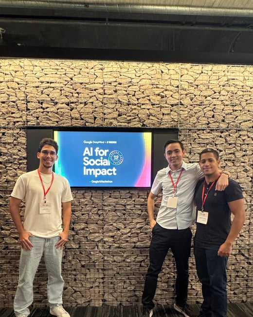 Three young men posing for a photo in front of a digital screen displaying 'AI for Social Impact' at a Google AI Hackathon event.
