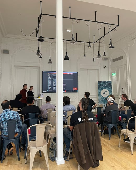 A photograph capturing an indoor technical presentation or meetup where two presenters stand beside a large screen displaying code, addressing a seated audience in a high-ceiling room with ornate white molding.