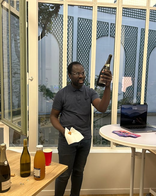 A photograph shows a man standing indoors, holding up a dark bottle in a celebratory manner, likely during an informal office gathering or social event. The scene is set against large, multi-paned windows, with tables nearby holding bottles, juice, and an open laptop displaying the time 19:49.
