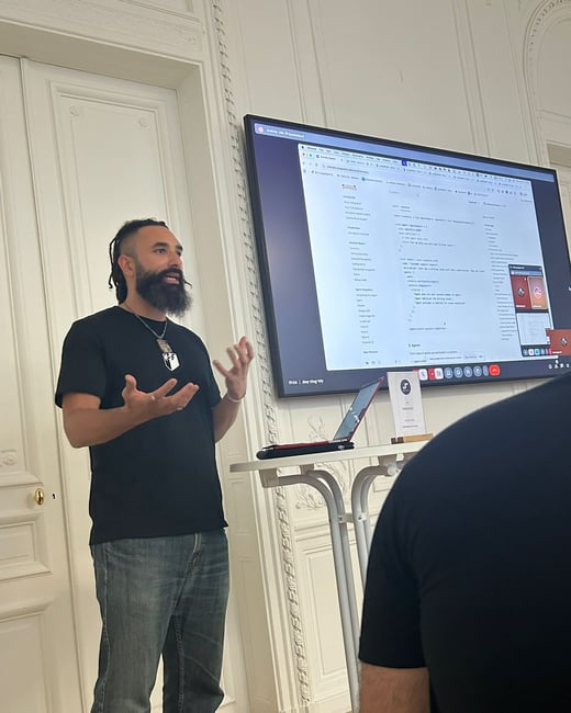 A man with a large beard and dreadlocks is giving a technical presentation, gesturing with his hands while standing next to a large screen displaying code and a table of contents.
