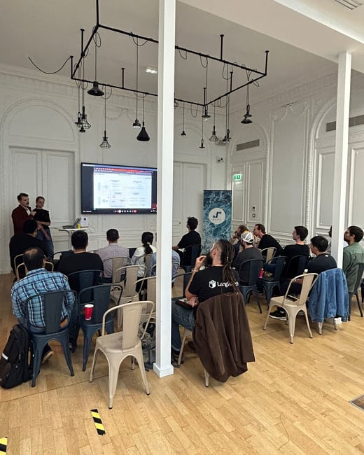 A candid photograph capturing an indoor presentation or tech talk, showing two speakers near a large screen displaying a diagram, and an audience seated in rows facing the front.