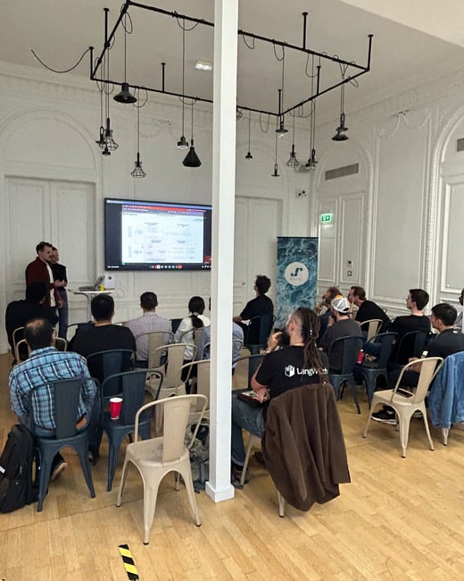 A photograph capturing an indoor presentation or technical talk, showing two speakers standing next to a large screen displaying diagrams, while an audience is seated facing them.