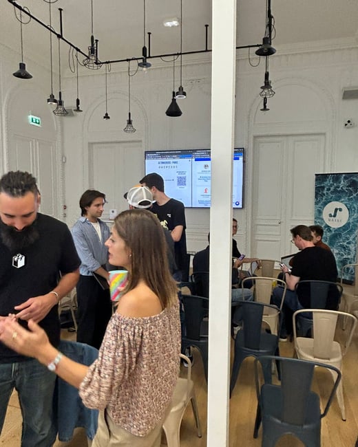 A candid photograph taken indoors at a professional or social gathering, showing several people networking or discussing in a modern event space with high ceilings and industrial lighting.