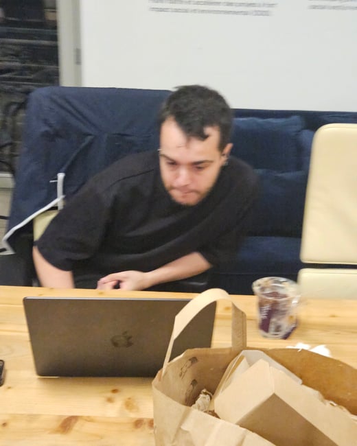 A candid photo of a young man sitting on a dark couch, looking down at a light wooden table where a closed laptop, a brown takeout bag, and a plastic cup are placed. The setting appears to be a casual indoor lounge or workspace.