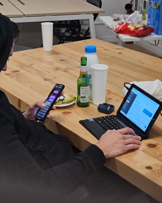 A person wearing a black hoodie sits at a wooden table, simultaneously using a smartphone and typing on a small laptop/tablet, surrounded by beverages and snacks.