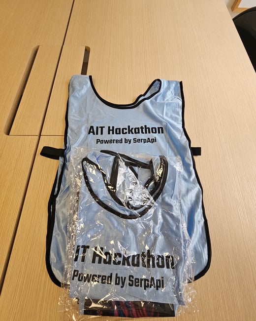 A photograph showing a light blue hackathon vest or bib, sponsored by SerpApi, lying flat on a light wood-grain table. The vest features black text and trim, and a portion of it is covered in clear plastic packaging.