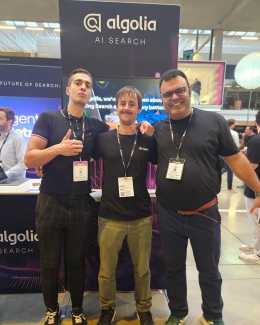 A candid photo showing three men posing together in front of a black Algolia booth, which prominently displays the company logo and the text 'AI SEARCH', likely taken at a professional conference or trade show.
