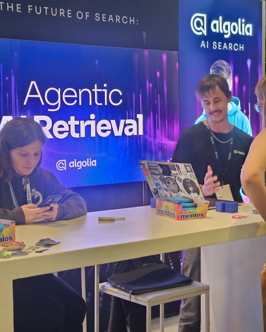 A candid photo capturing three people interacting at a high table in a conference booth, featuring large screens displaying the text "Agentic Retrieval" and Algolia branding.