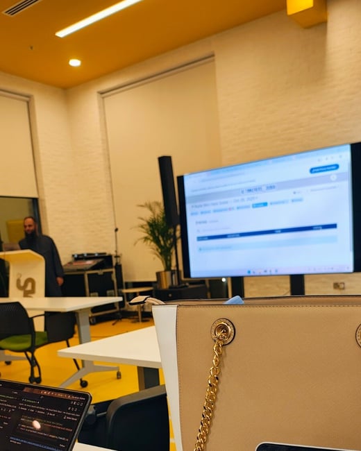 A candid indoor photograph capturing a technical workshop or presentation from the perspective of an attendee, showing laptops, handwritten notes, and a stylish handbag in the foreground, with a presenter standing at a podium and a large screen displaying information in the background.