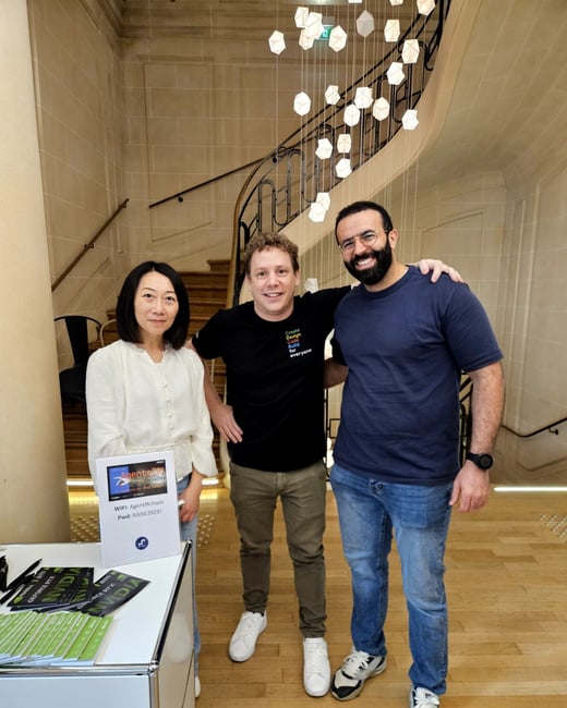 A photo showing three people (one woman and two men) posing together in the lobby of a grand building, likely at an event, with a large curved staircase and a modern geometric chandelier visible behind them.