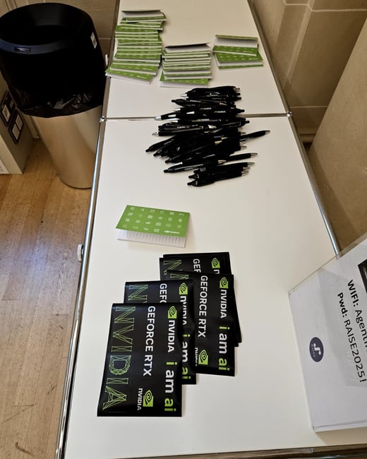A photograph showing a white table covered with promotional merchandise, including piles of black NVIDIA pens, green notepads, and black stickers featuring NVIDIA, GeForce RTX, and 'i am ai' branding.