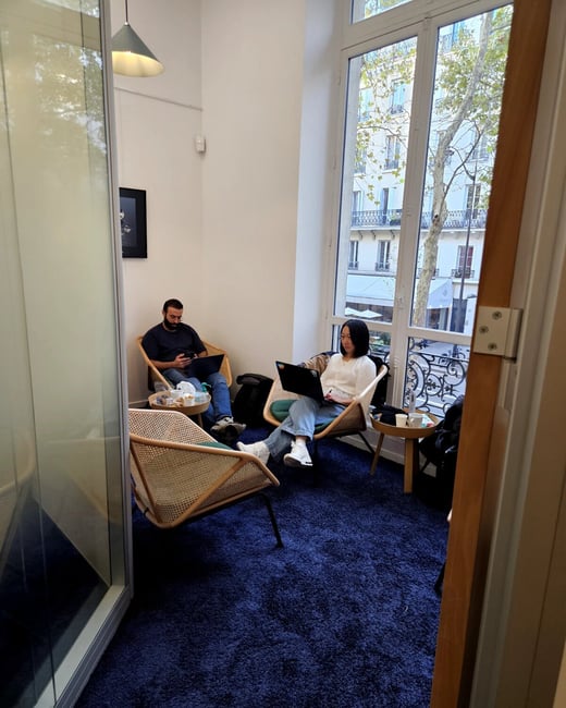 A candid photograph shows two individuals, a man and a woman, sitting in modern wicker chairs in a bright room with a deep blue shag carpet, both working on open laptops. The room has a large window offering a view of a classic European urban street scene outside.