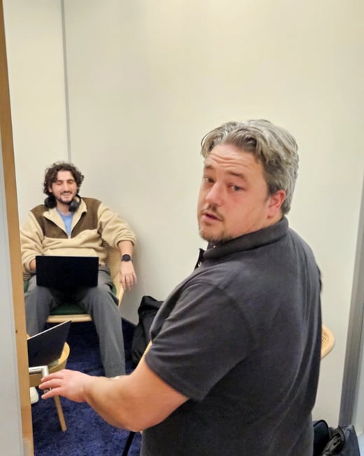 A candid indoor photograph showing two men; one man in the foreground, wearing a dark polo shirt, is looking back toward the camera, while another man is seated in the background with a laptop on his lap.