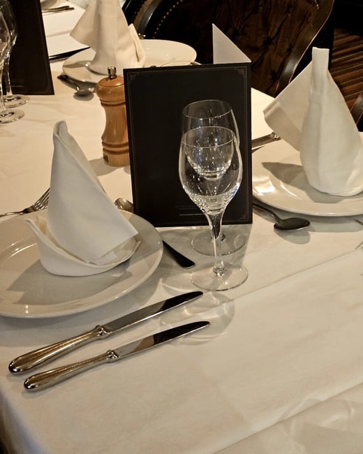 A close-up photograph of a formally set dining table in a restaurant, featuring white linen, plates, neatly folded white napkins, silverware, and two stacked wine glasses.