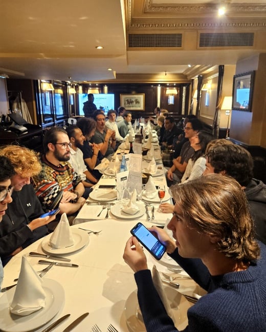 A large group of people is seated around a long, formally set table inside an upscale restaurant with dark wood paneling and ornate ceilings, engaged in dining and conversation.