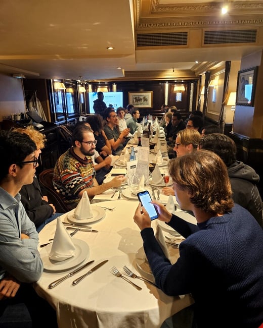 A candid photograph captures a large group of people seated around a long, oval table in a dimly lit, ornate restaurant, engaged in a dinner gathering while a presentation screen is visible in the background.