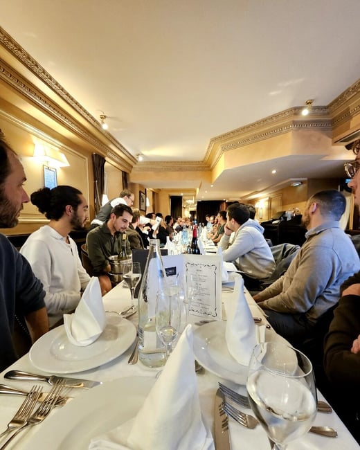 A candid photograph captures a large group of people seated around a long, formally set dining table inside a warmly lit restaurant with ornate, golden-hued decor. The foreground shows detailed place settings with white plates, silverware, and tall folded white napkins.