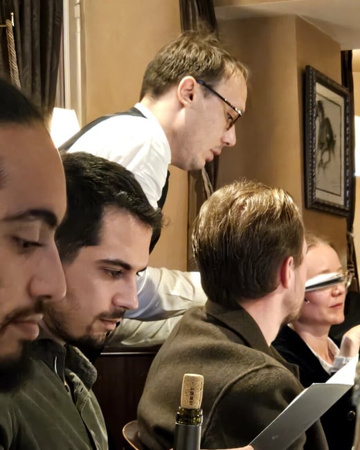 A candid indoor photograph captures a group of people seated at a restaurant table, with a waiter leaning over them, seemingly taking an order or discussing the menu. A wine bottle with a cork is prominent in the foreground.