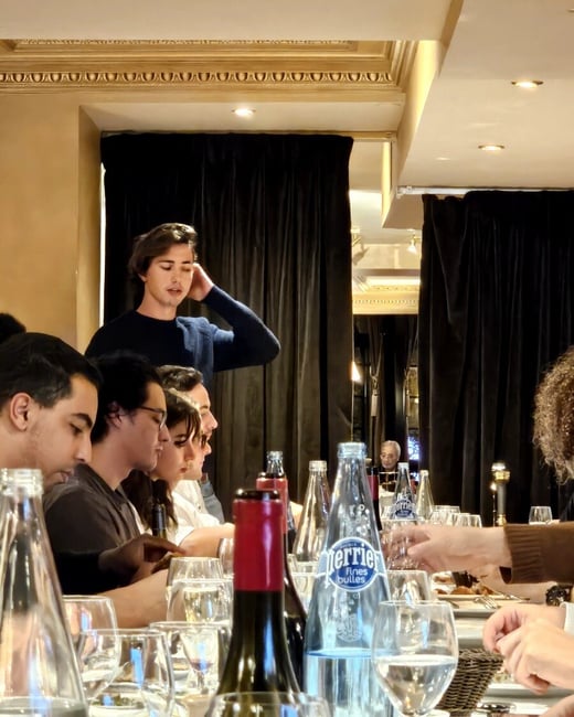A candid photograph captures a group of people seated at a long dining table in an ornate restaurant setting, characterized by dark curtains and gold molding, with a young man standing up and looking down at the group.