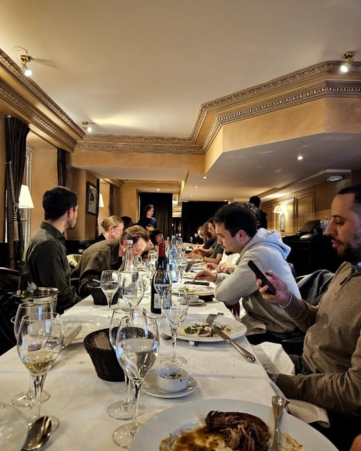 A candid photograph captures a group of people seated at a long table enjoying a dinner in an upscale restaurant with ornate, warm-toned interior decor.