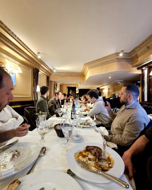 A candid photograph captures a large group of people seated at a long table in a formal, ornately decorated restaurant, engaged in a social dinner.