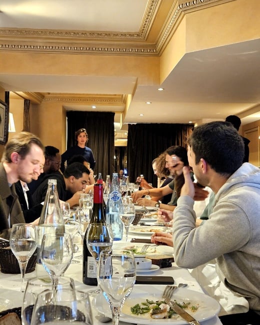 A candid photograph captures a group of people dining together at a long, formal table in a restaurant with ornate golden walls and crown molding. The table is heavily set with wine glasses, bottles, and plates of food.