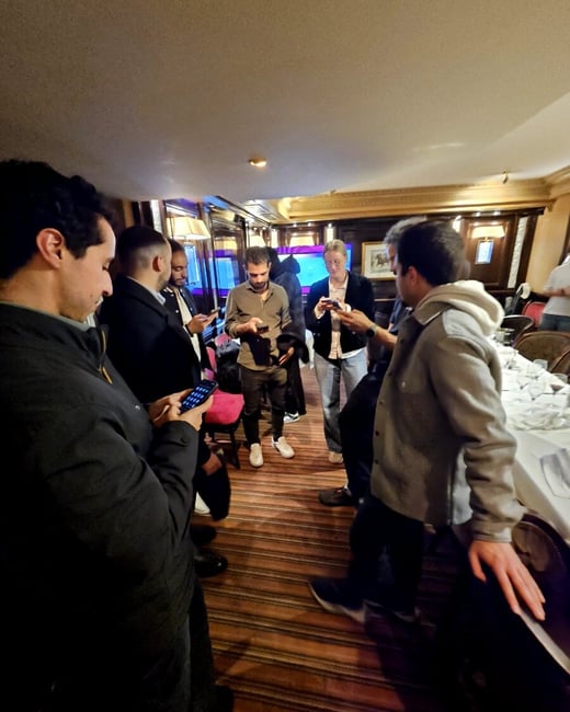 A candid photograph captures a small group of people gathered in a dimly lit, ornate indoor space, such as a restaurant or lounge, with most individuals looking down at their smartphones.
