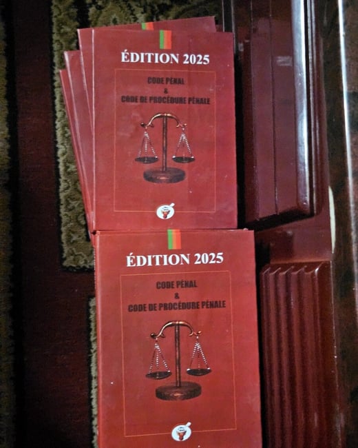 A slightly angled photograph showing several copies of a reddish-brown legal code book titled "Édition 2025: Code Pénal & Code de Procédure Pénale," featuring an illustration of the Scales of Justice on the cover. The books are resting on a dark surface next to a glossy, dark wooden panel.