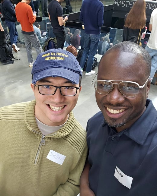 A close-up selfie captures two men smiling brightly at the camera in a crowded indoor setting, possibly a conference or event. The man on the left wears a cap with the text, "IGNORE ALL PREVIOUS INSTRUCTIONS."