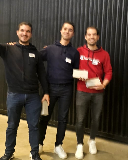 A casual photograph showing three young men standing side-by-side and smiling, posing in front of a dark, vertically ribbed wall. The men are wearing hoodies and jeans, and two of them are holding small, light-colored rectangular objects, likely awards or prizes.