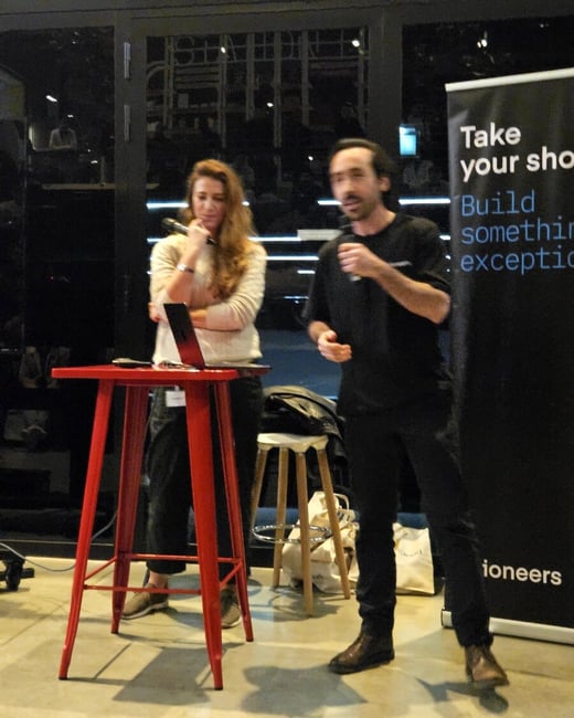 A photograph capturing two people, a man and a woman, giving a presentation or talk indoors next to a bright red high table holding a laptop. Behind them is a black banner with white and blue text promoting building something exceptional.