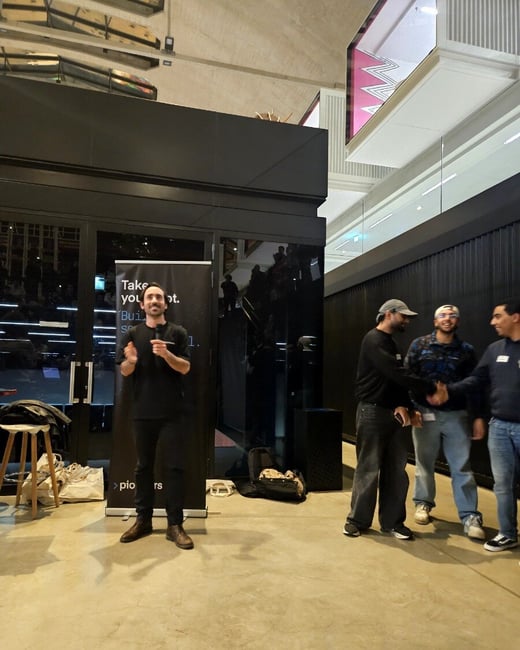 A photograph taken indoors at a modern event space, showing a man presenting in front of a dark banner while three other male attendees stand to the right, two of whom are shaking hands.