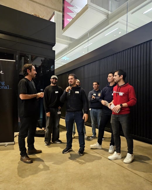 A group of seven men is gathered in a modern indoor venue, where one man in the center is speaking into a microphone while others listen, suggesting an informal presentation or discussion.