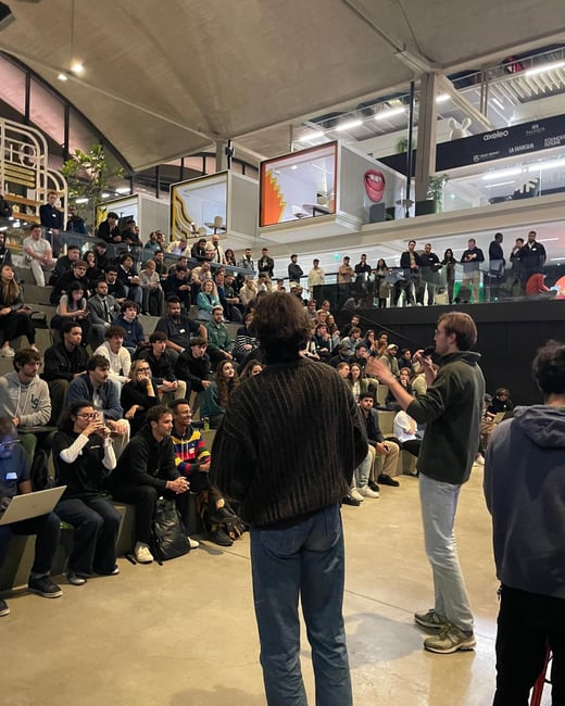A large crowd of people is seated in a modern, tiered indoor amphitheater, watching two individuals who are standing in the foreground and appearing to lead a discussion or presentation. The venue features industrial architecture and visible company logos on an upper balcony.