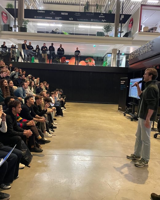 A photograph capturing an indoor presentation event where a speaker, holding a microphone, addresses a large seated audience in a modern, multi-level architectural space featuring corporate sponsor logos on a banner above.