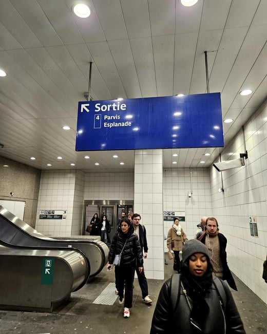 A candid photograph taken inside a brightly lit subway or metro station, showing several commuters walking past an escalator and beneath a large blue directional sign pointing toward 'Sortie' (Exit).