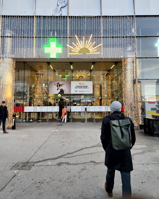 A photograph captures the glass entrance facade of a large Westfield shopping center decorated with numerous white string lights, featuring a prominent illuminated green cross and a golden sunburst motif. A person wearing a dark coat and a green backpack stands in the foreground with their back to the camera, looking toward the entrance.