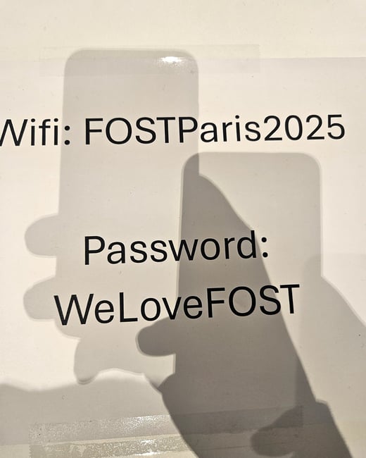 A close-up photograph displays the WiFi network name "FOSTParis2025" and the password "WeLoveFOST" printed in black text on a white surface, partially covered by the dark shadows of two mobile phones and a hand.