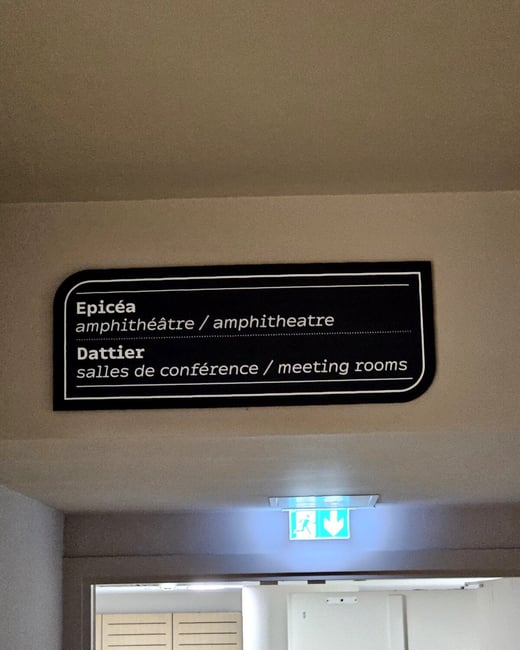 A photograph showing a black directional sign mounted on a beige wall in an indoor hallway, indicating 'amphithéâtre / amphitheatre' and 'salles de conférence / meeting rooms'.