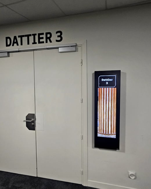 A photograph showing the entrance to an indoor room labeled 'DATTIER 3' in large black text above white double doors, next to a vertical digital display screen repeating the room name and showing a striped graphic.