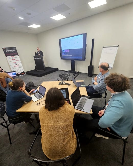 A photograph capturing an indoor technical presentation or workshop where a speaker stands on a stage behind a podium, addressing a small group of attendees seated around a hexagonal table with open laptops.