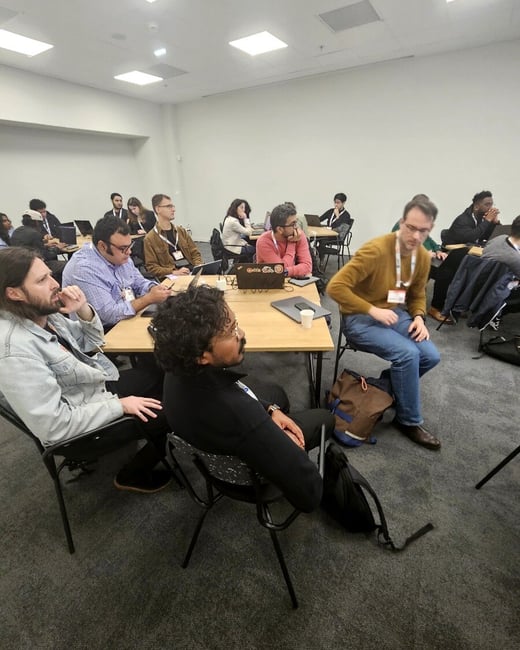 A candid photograph captures a group of people seated at wooden tables in a brightly lit indoor conference room or workshop, attentively facing a presentation area outside the frame. Many attendees have laptops, notebooks, and visible conference badges.
