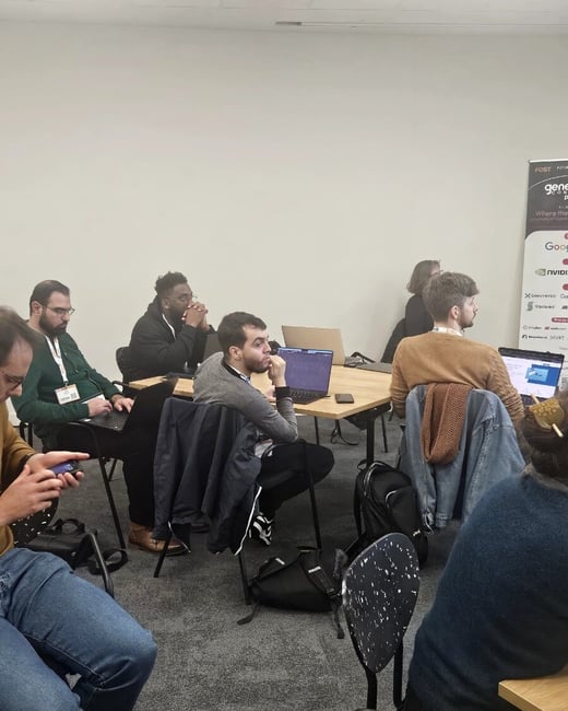 A photograph showing seven attendees seated around a wooden table in a brightly lit indoor space, focusing on their laptops or listening during what appears to be a tech workshop or conference session.