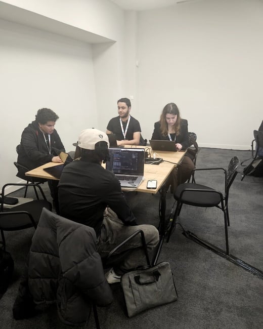A candid photograph shows four individuals seated around a wooden table in a simple, brightly lit room with white walls and dark carpet, engaged in collaborative work on their laptops.