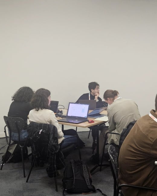 A candid indoor photograph shows five people seated around a square table, primarily focused on working on their open laptops in what appears to be a conference or workshop setting with a plain white wall background.