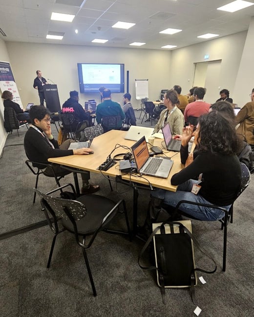 A photograph capturing an indoor technical workshop or conference session, showing attendees seated at tables with laptops, listening to a presenter standing at a podium near a large projection screen displaying data.