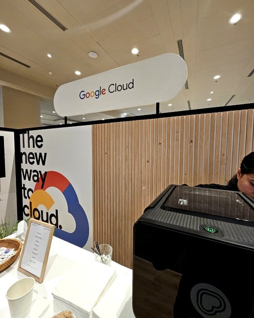 A candid photograph of an indoor event booth sponsored by Google Cloud, featuring a service counter, branding signage, and a large black machine (likely for beverage service) being attended by a person.