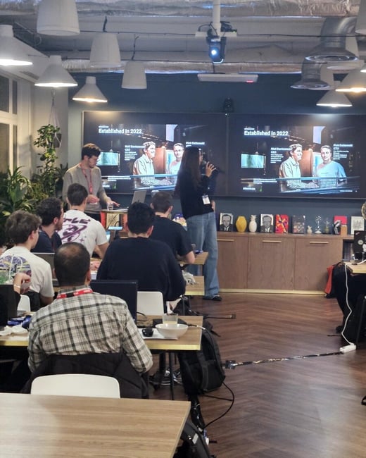 A photograph captures an indoor presentation or workshop where attendees are seated at tables facing a large display screen that shows information about two company executives. A woman stands near the screen, speaking into a microphone, while a man stands beside her.