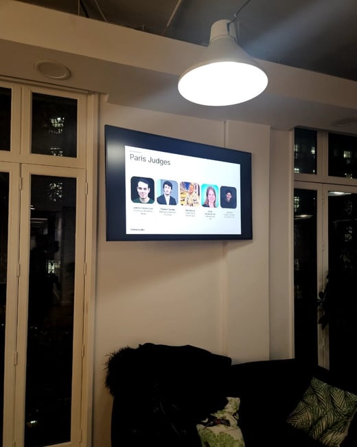 A low-light photograph of an indoor space showing a wall-mounted television screen displaying a presentation slide titled "Paris Judges" with five headshots and corresponding names. The scene includes a bright overhead pendant light, white walls, large glass doors, and a dark sofa with pillows in the foreground.